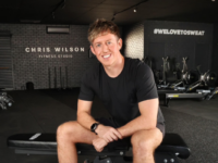 Man in black gym clothes sitting in dark gym with fitness machines in the background