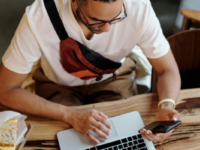 Man with a laptop and sandwich on the desk in front of him using a phone.