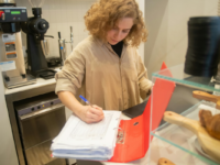 A cafe worker in a cafe kitchen writes on a piece of paper in a binder.