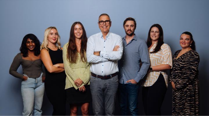 A group of people of mixed genders and ages standing against a grey wall.