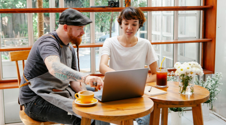 A woman and a man in an apron work on a laptop at a cafe table.