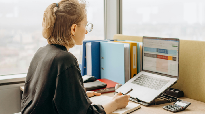 Woman sitting at desk looking at laptop with spreadsheet application open.