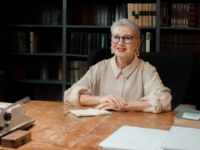 A woman sitting at a desk in front of a bookshelf with her arms folded.