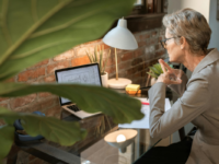 A woman works on a laptop at a desk with a houseplant in the foreground.