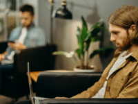 Man in brown jacket using laptop in cafe with other worker behind him.